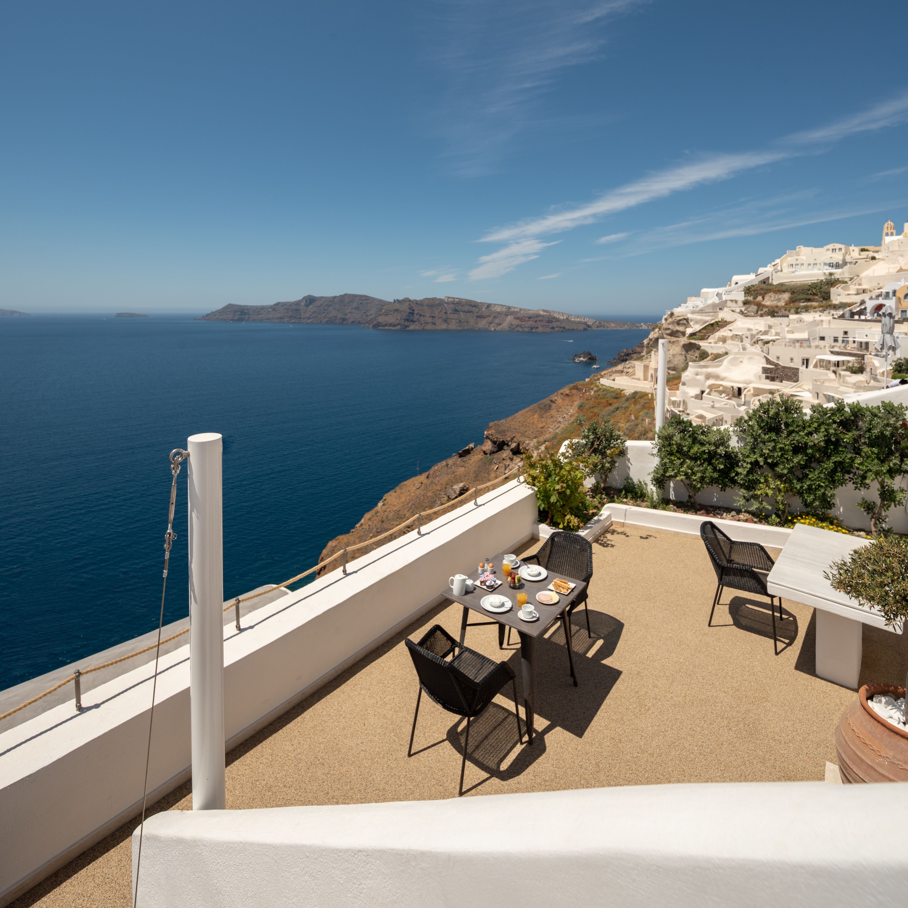 A terrace with a table set for dining, overlooking a coastal landscape with buildings and the sea.