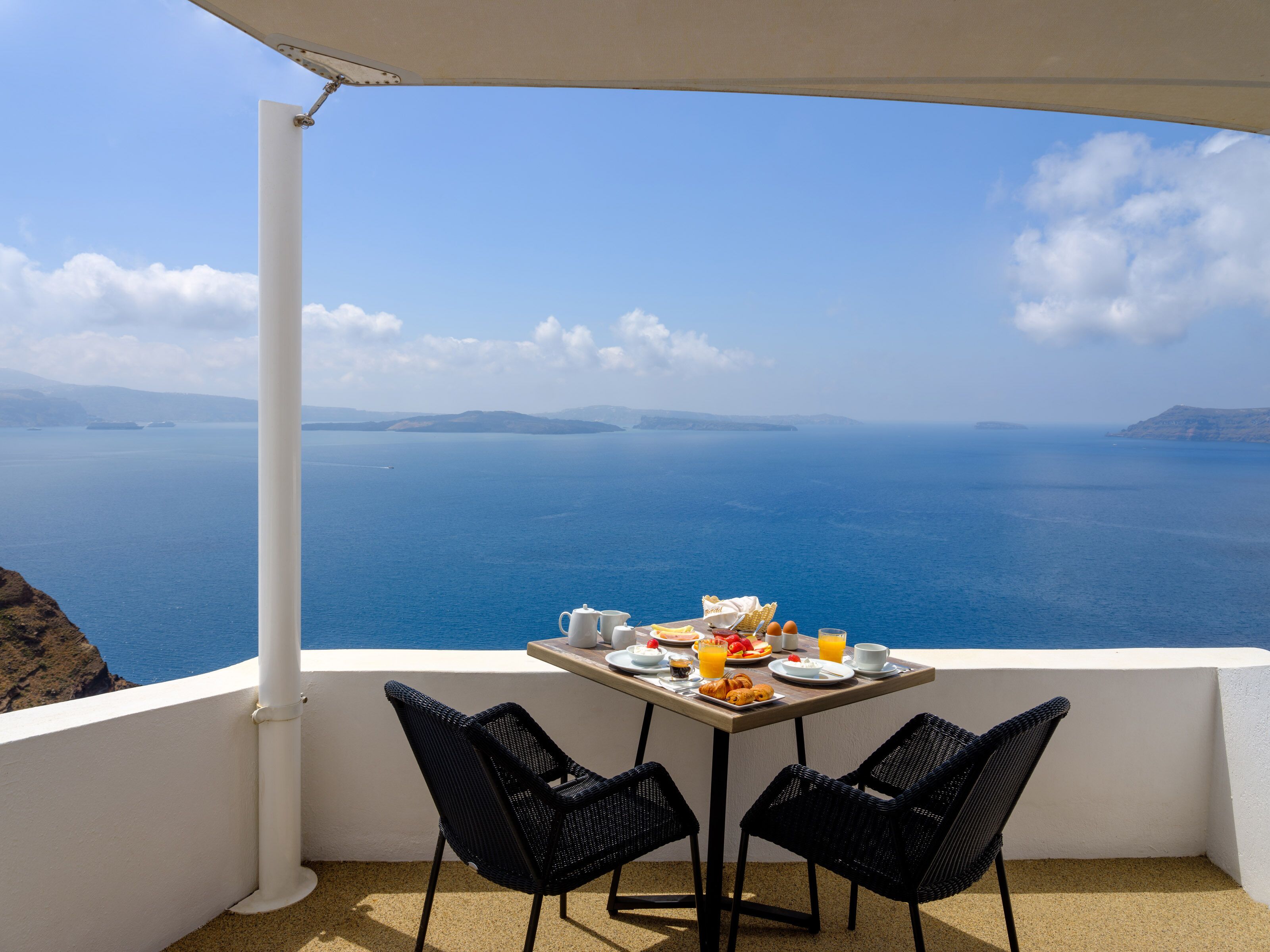 A balcony with a table set for breakfast overlooking a scenic view of the sea and distant islands.