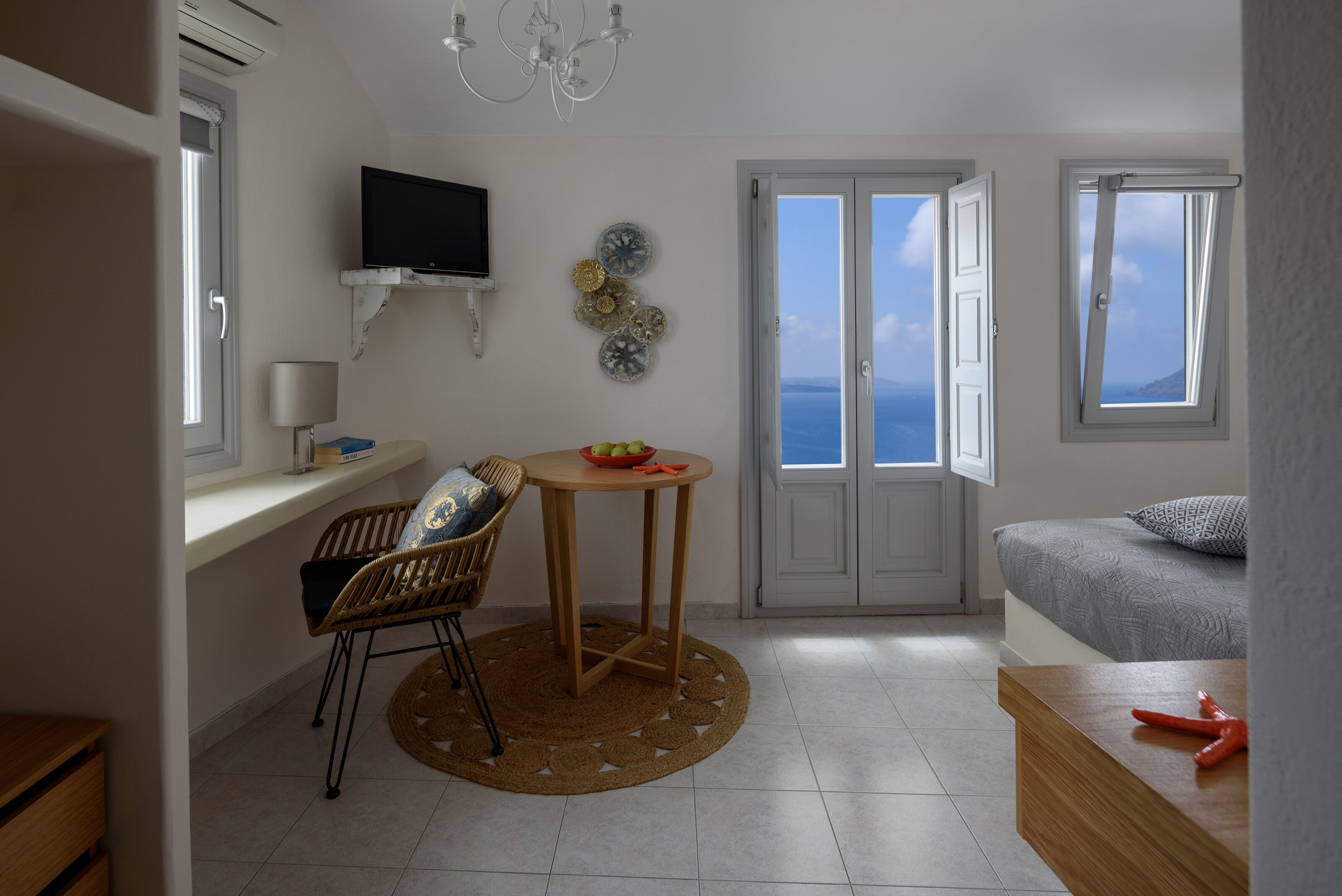 Un espacio habitable pequeño con una cama, una mesa con un frutero, una silla, un televisor montado en la pared y vistas al mar a través de una puerta.