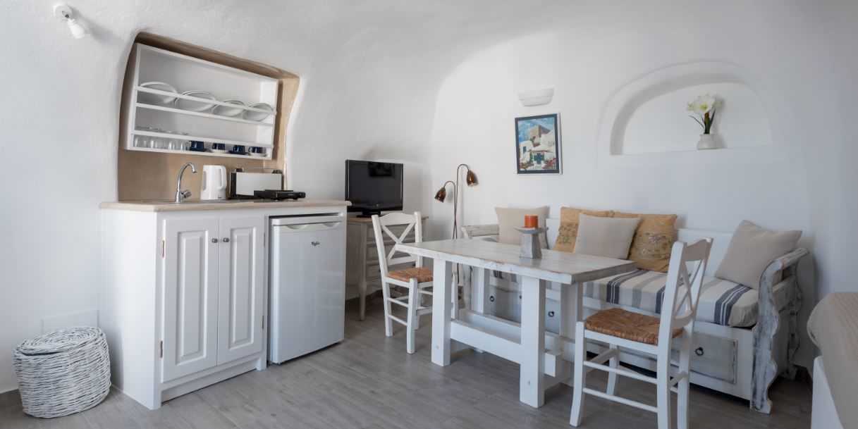 A compact, white-themed room with a kitchenette, a dining area with a table and chairs, and a sofa with cushions.