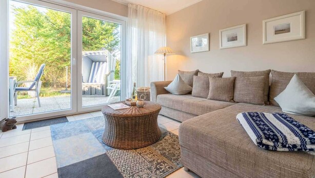 Ein Wohnzimmer mit einer Couch, einem runden Couchtisch und einer Lampe. An der Wand befinden sich gerahmte Bilder und ein Blick ins Freie durch eine Schiebetür.