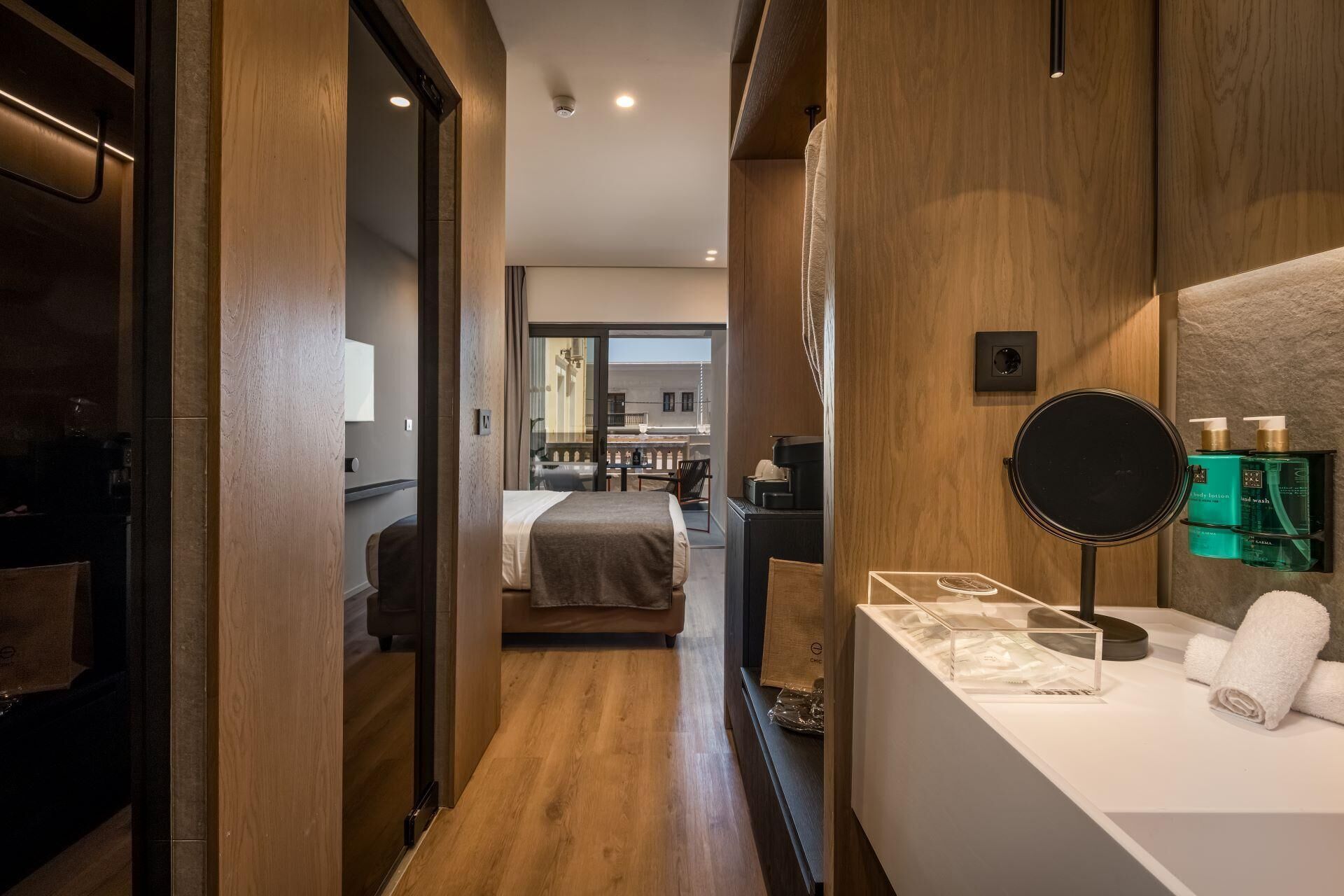 Ein modernes Hotelzimmer mit Holzboden, einem Bett, einem Badezimmer mit Spiegel und Pflegeprodukten sowie einem Blick auf den Balkon.