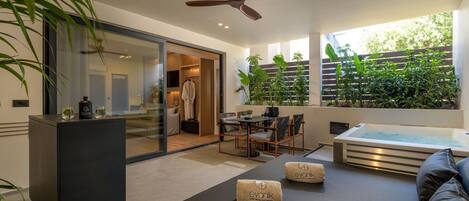 A modern hotel room with a sliding glass door leading to a balcony, a ceiling fan, a seating area with a table and chairs, and a hot tub.