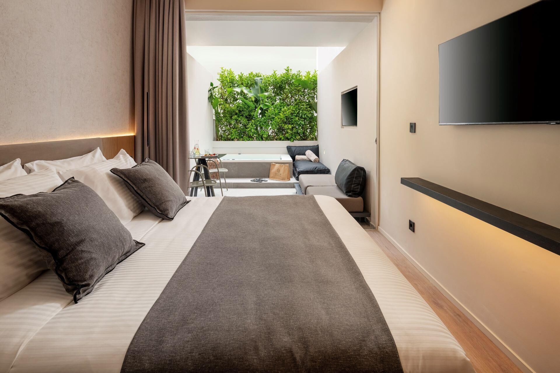 Ein modernes Hotelzimmer mit einem großen Bett, einem an der Wand befestigten Flachbildfernseher und Blick auf einen üppigen, grünen Garten durch einen offenen Balkon.