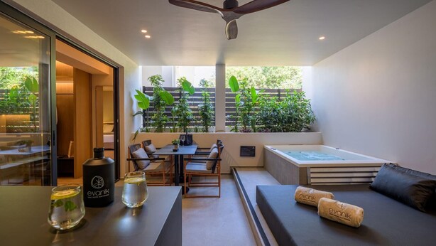 A modern hotel room with a glass wall, a dining area, a seating area with a sofa and cushions, a hot tub, and a ceiling fan. - Evanik Chic Hotel (Kalymnos)