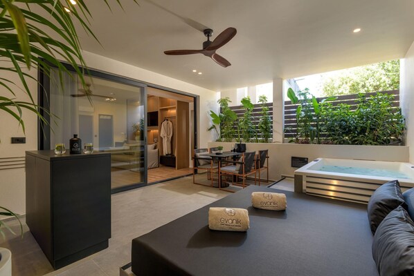 A modern room with a black sofa, a table with towels, a ceiling fan, a glass door leading to a bathroom, and a large window with plants. - Evanik Chic Hotel (Kalymnos)