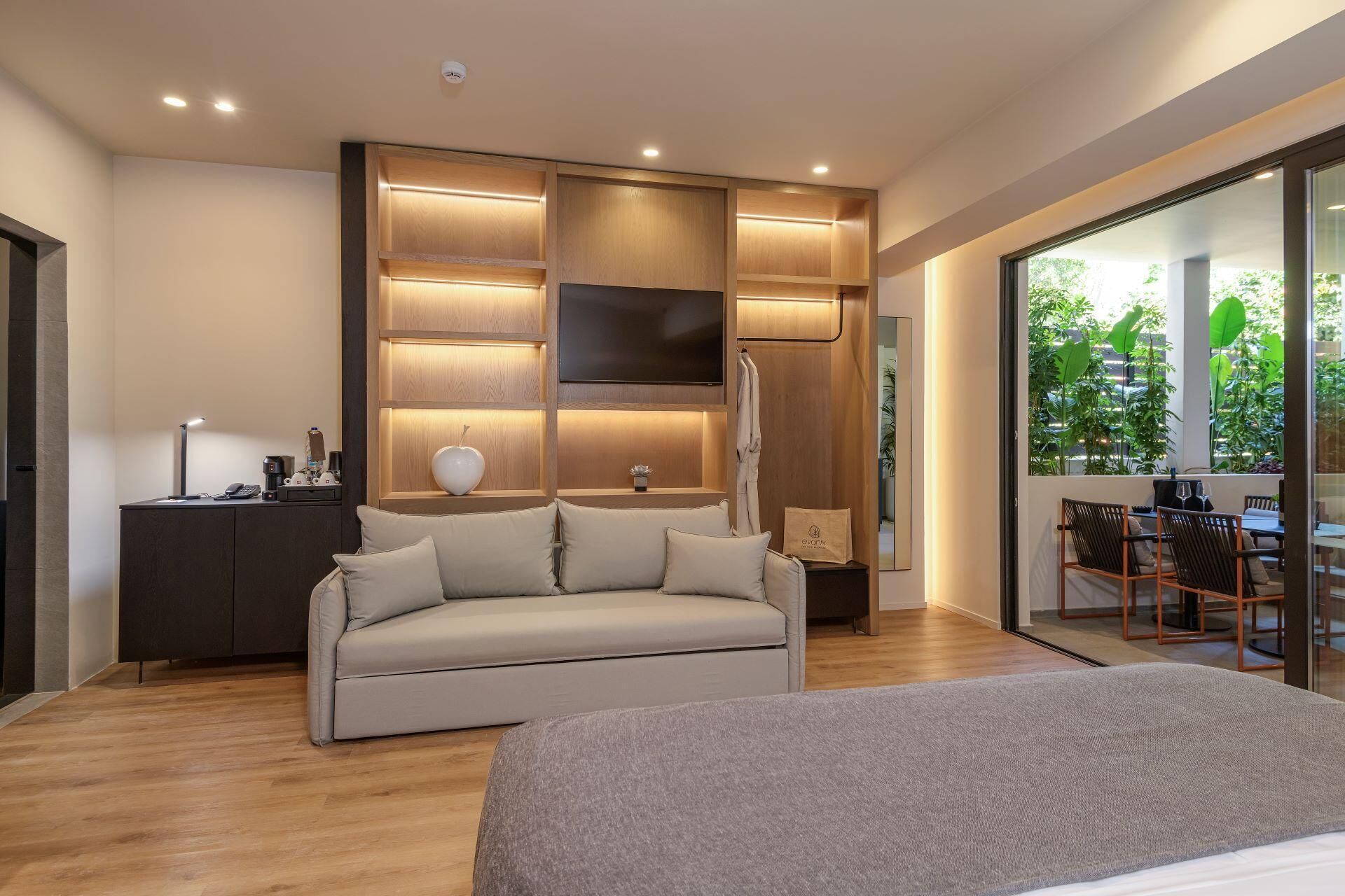 A modern hotel room with a sofa, a bedside table, a TV, and a large window with a view of greenery.
