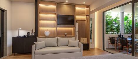 A modern hotel room with a sofa, a bedside table, a TV, and a large window with a view of greenery.