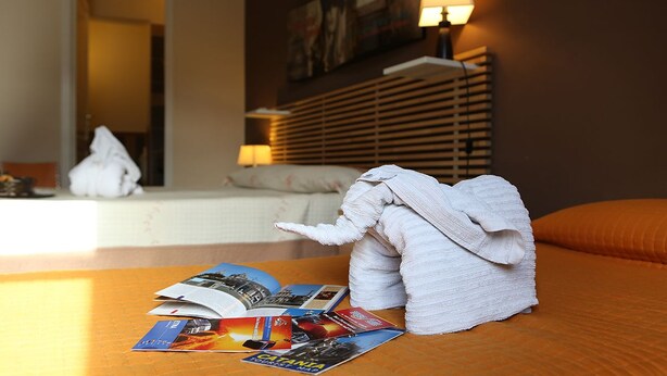 A neatly made bed with a folded towel, a lamp, and brochures on a wooden table in a hotel room. - Le 3 B (Catania)