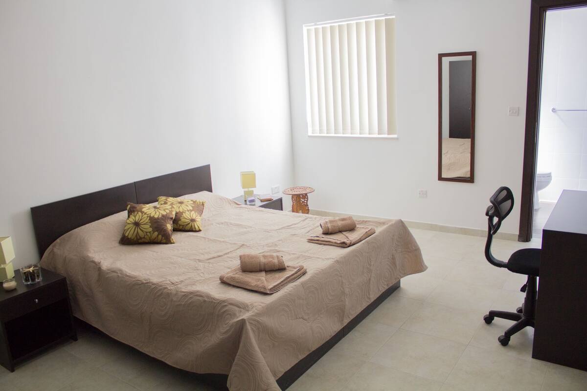 Quarto bem arrumado com uma cama grande, uma mesa de cabeceira, uma cadeira, uma mesinha e uma janela com persianas.