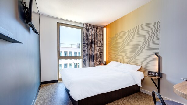 Una habitación de hotel con una cama grande, un escritorio con silla, una ventana con cortinas y una lámpara de pared.