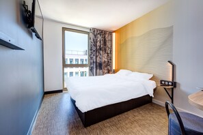 Ein Hotelzimmer mit einem großen Bett, einem Schreibtisch mit Stuhl, einem Fenster mit Vorhängen und einer Wandleuchte.