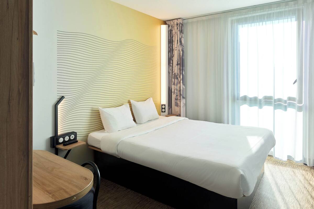 Habitación de hotel con una cama grande, una mesita de noche con lámpara, una pequeña mesa y vistas al exterior a través de cortinas traslúcidas.