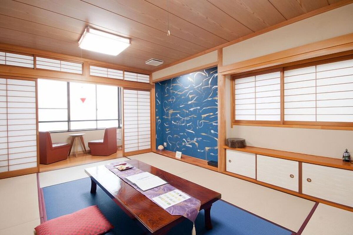 A traditional Japanese room with tatami flooring, a low table, sliding doors, and a view of the sky through the windows.