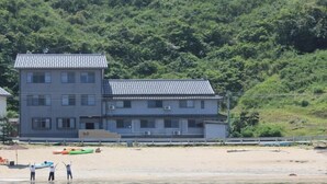 A building with a tiled roof, a sandy beach, and a forested hillside. - Yuyado Umi no Ne (Toyooka)