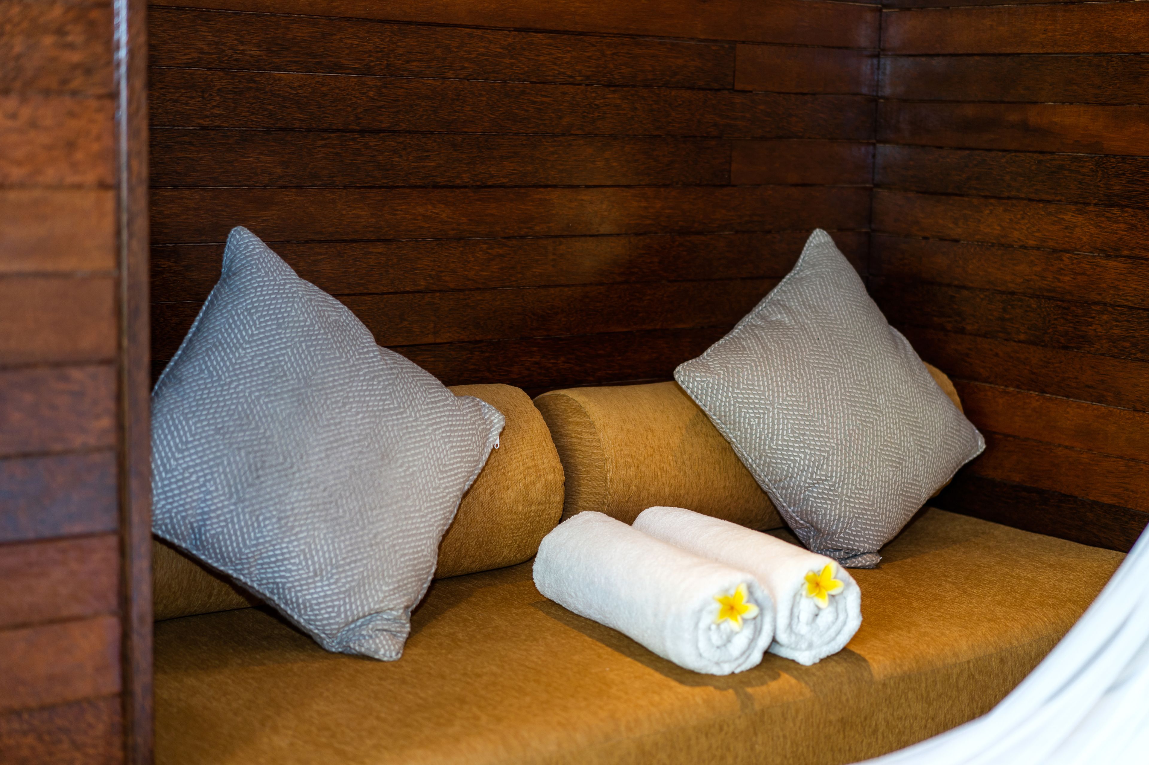 A wooden cabin-style room with a brown sofa, two grey pillows, and two rolled towels with yellow flowers.
