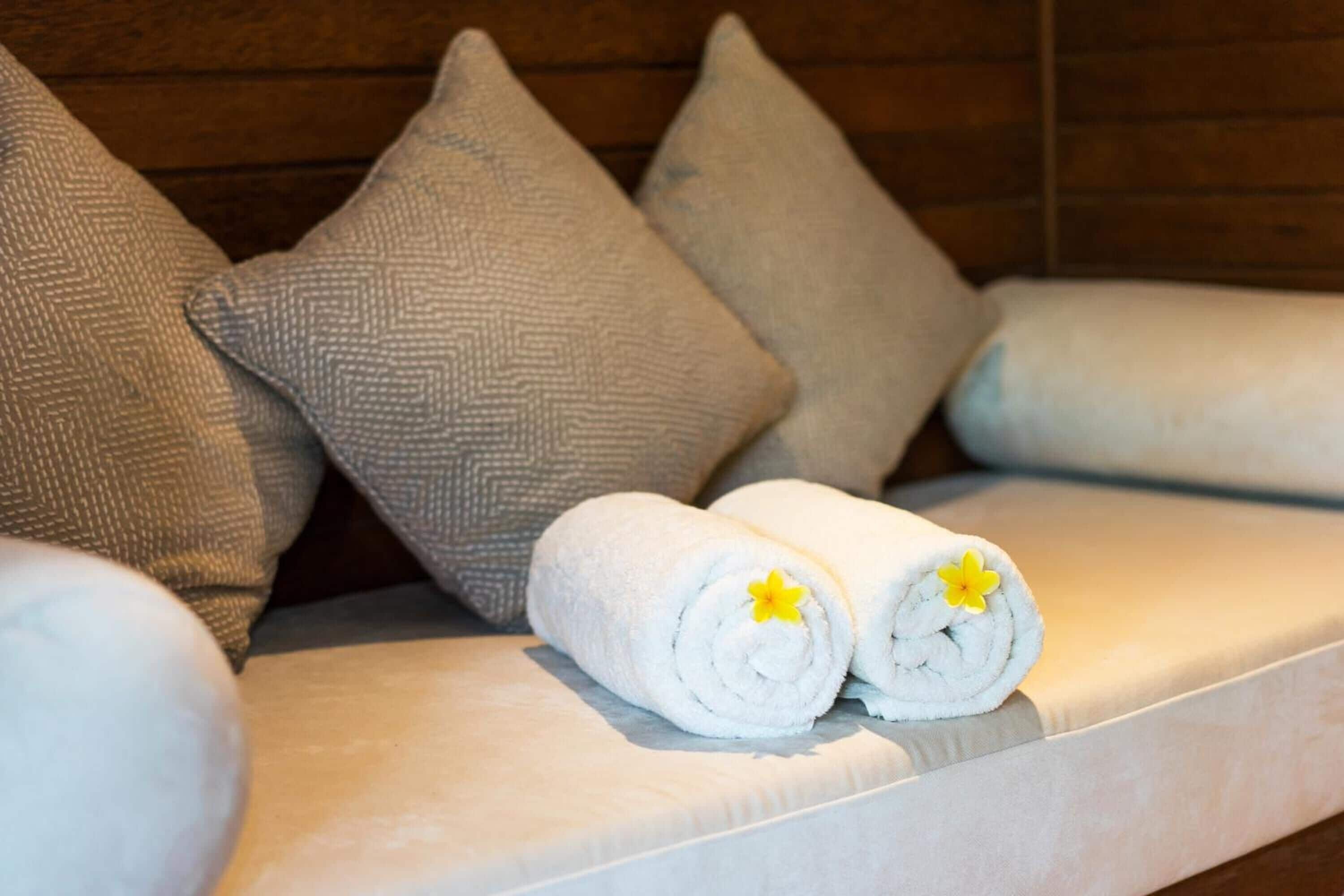 A sofa with rolled white towels and a yellow flower, set against a wooden wall.