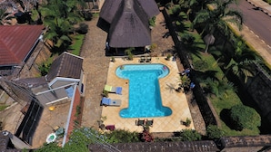 A large swimming pool with a unique shape, surrounded by palm trees and a thatched-roof structure. - Entebbe Palm Hotel (Entebbe)