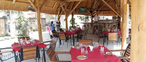 Uma área de refeições ao ar livre com mesas preparadas para dois, coberta por uma estrutura de telhado de madeira.