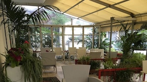 A covered outdoor seating area with wicker furniture, potted plants, and a view of the surrounding greenery. - Noi Due Hotel (Rimini)