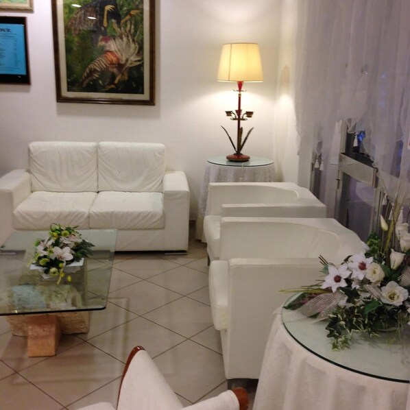 Una sala de estar con un sofá blanco, una mesa de centro de vidrio y una lámpara. Hay un cuadro en la pared y un arreglo floral sobre la mesa.
