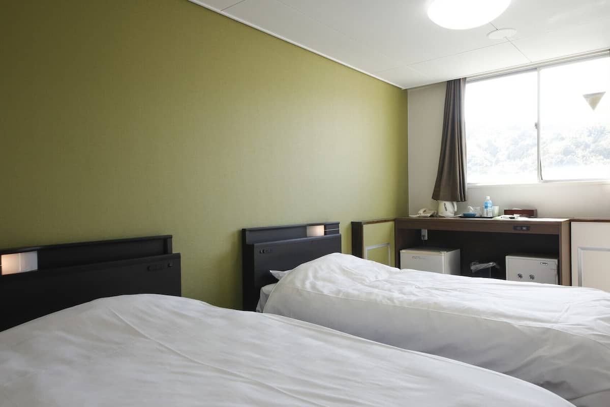 Ein Hotelzimmer mit zwei Betten, einem Schreibtisch, einem Fenster mit Vorhängen und einer kleinen Küchenzeile.