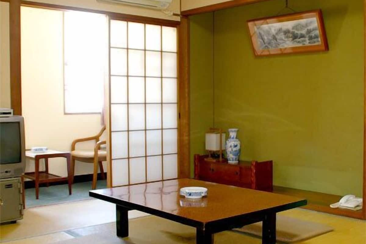 Ein traditioneller japanischer Raum mit Tatami-Boden, einem niedrigen Tisch, einem Fernseher und einer Schiebetür.