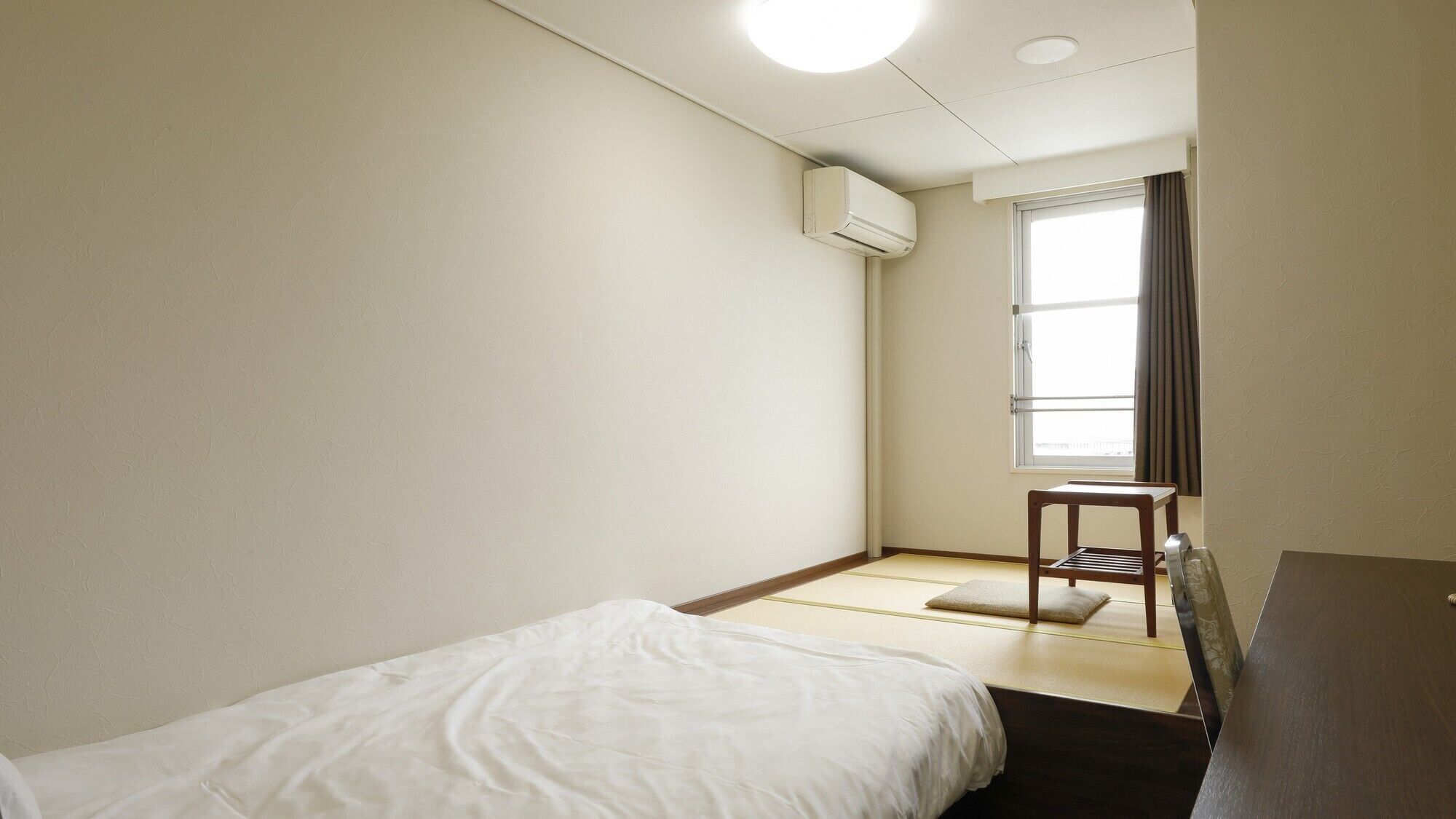 A small, clean room with a bed, a chair, a window, and an air conditioning unit.