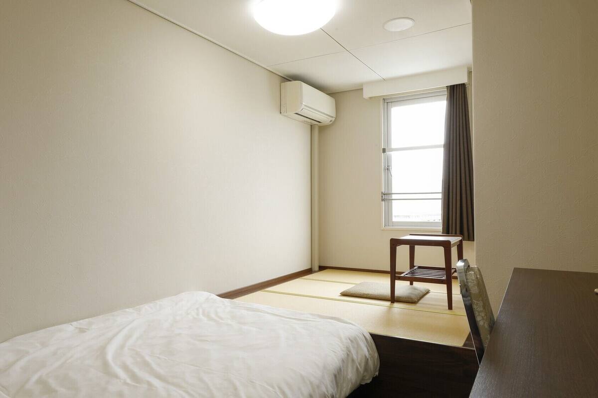 Ein kleines, sauberes Zimmer mit Bett, Stuhl, Fenster und Klimaanlage.