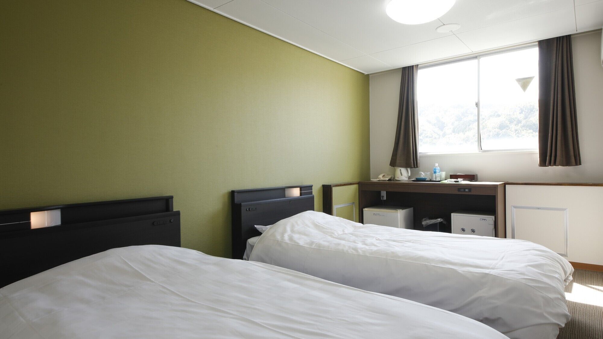 A hotel room with two beds, a desk, a window with curtains, and a small kitchenette.