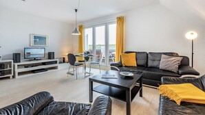 A modern living room with a black leather sofa, a coffee table, and a TV on a white stand. There is a dining area with a table and chairs, and a balcony with yellow curtains. - Apartment 5 (ID 171), Germany (Sankt Peter-Ording)