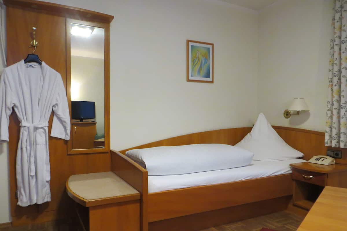 A hotel room with a wooden bed, a bedside table with a lamp, a telephone, a robe hanging on a rack, and a painting on the wall.