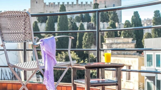 A balcony with a metal chair, a small table, and a glass of orange juice. In the background, there is a castle-like structure. - Danezis City Stars (Rhodes)