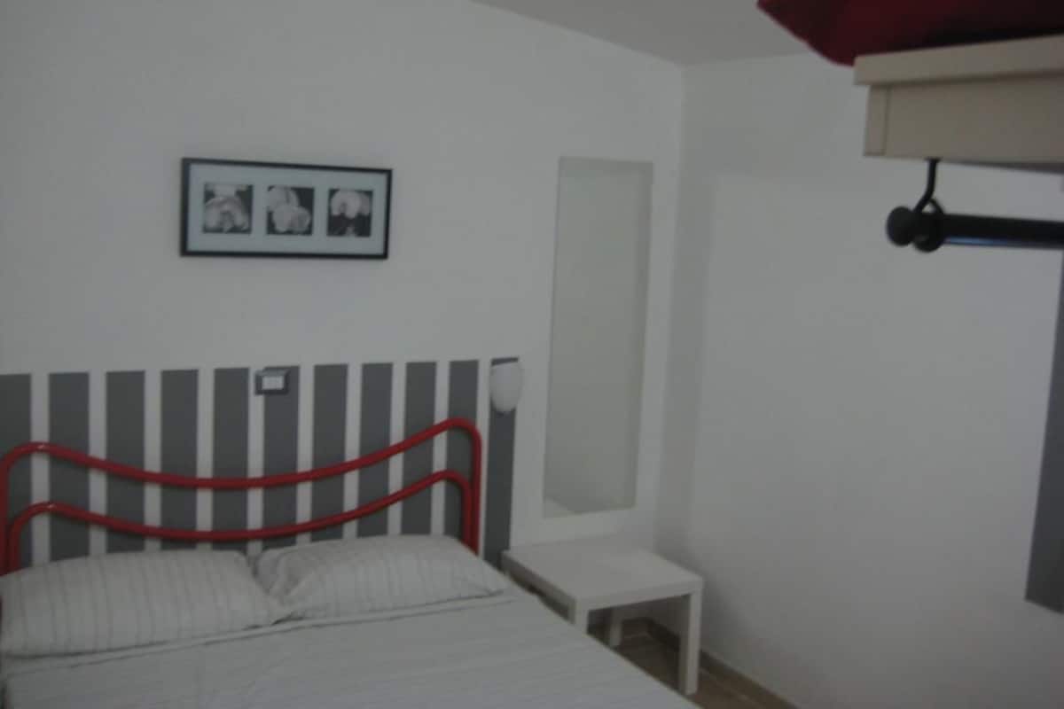 Une chambre à coucher avec un lit, une table de chevet, une commode, une tête de lit rouge, un tableau encadré au mur et une fenêtre avec un store blanc.