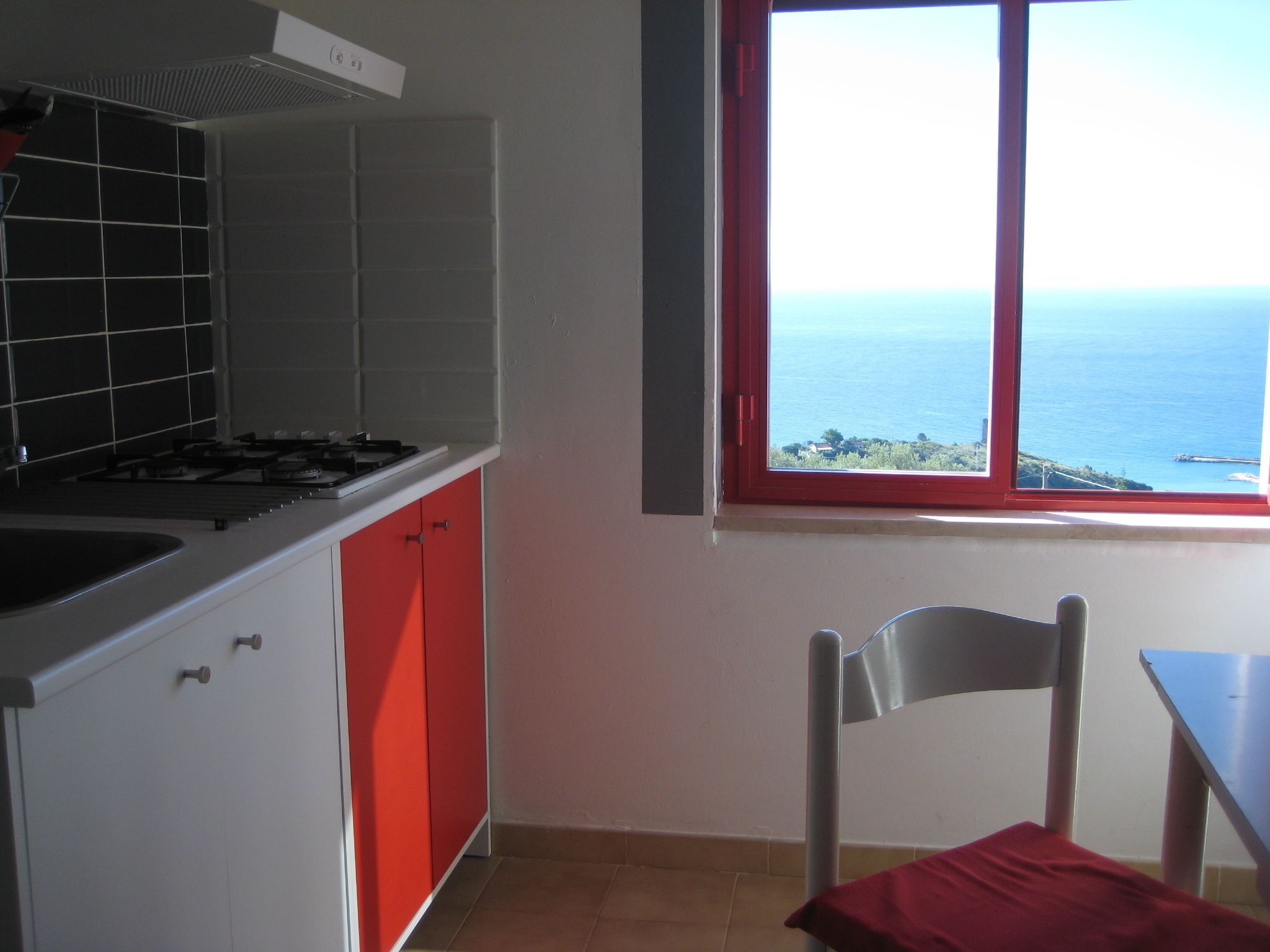 A kitchen with a gas stove, red cabinets, and a window overlooking the sea.