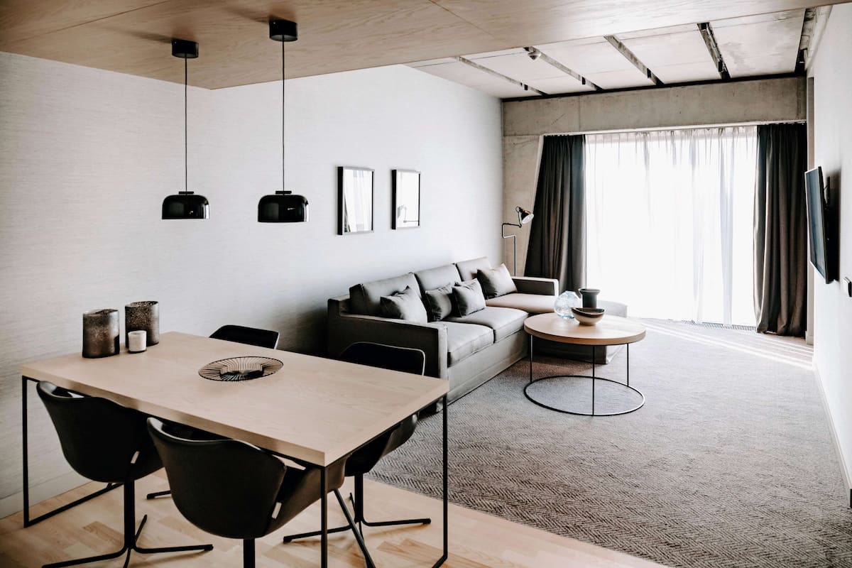 Un salon moderne avec une table à manger en bois, des chaises noires, un canapé gris et une table basse ronde. On y trouve deux suspensions noires, un lampadaire et un téléviseur fixé au mur.