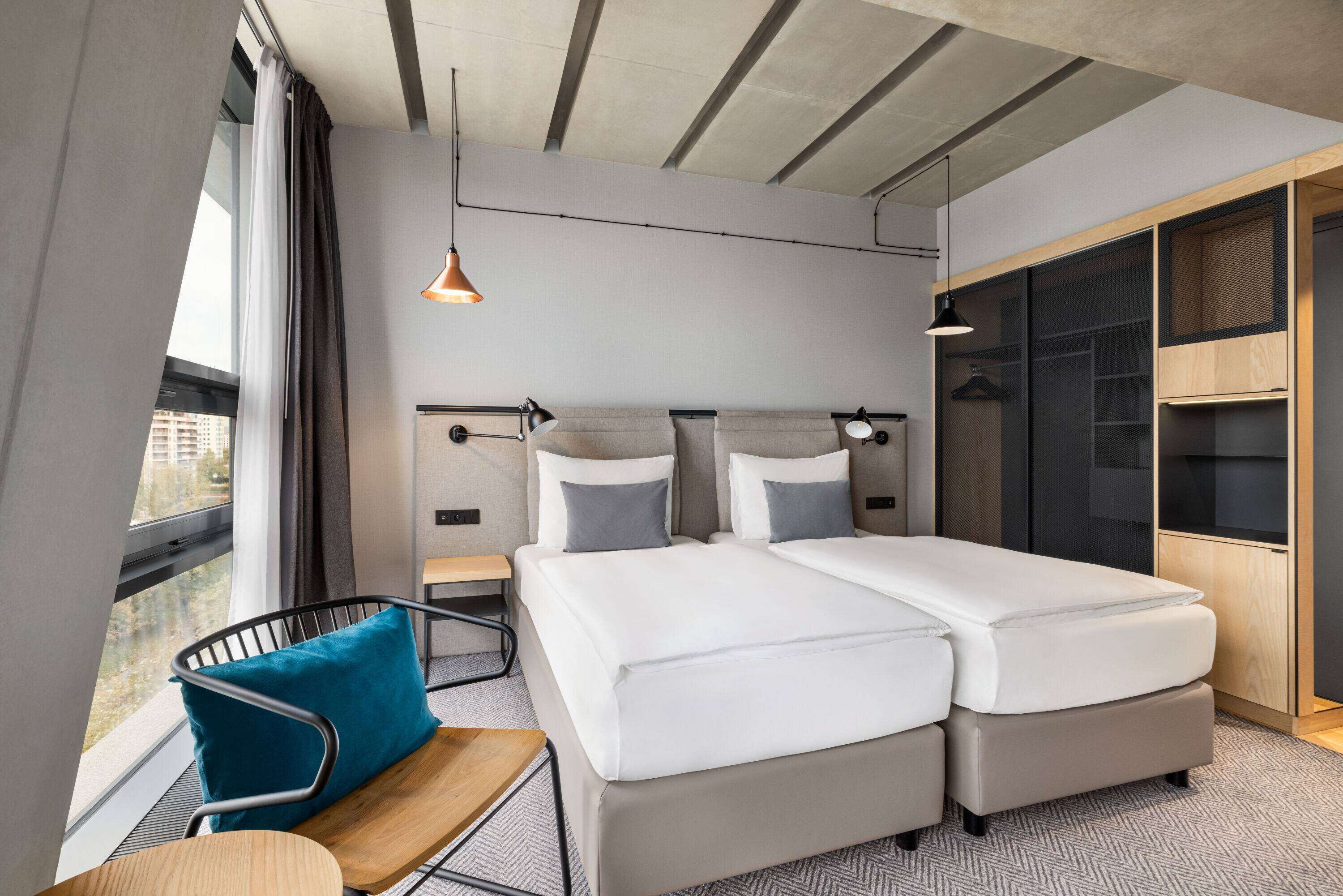 A modern hotel room with a double bed, a desk with a chair, a wardrobe, and a large window with curtains.