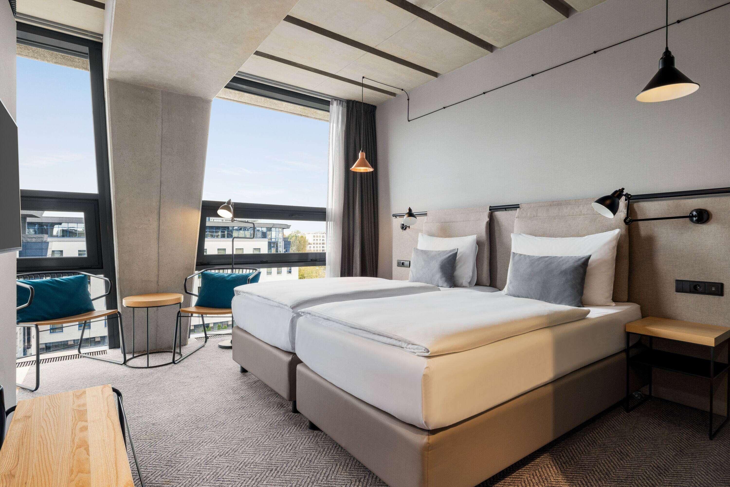 A hotel room with a large bed, a desk, a chair, a small table, and a view of the city.
