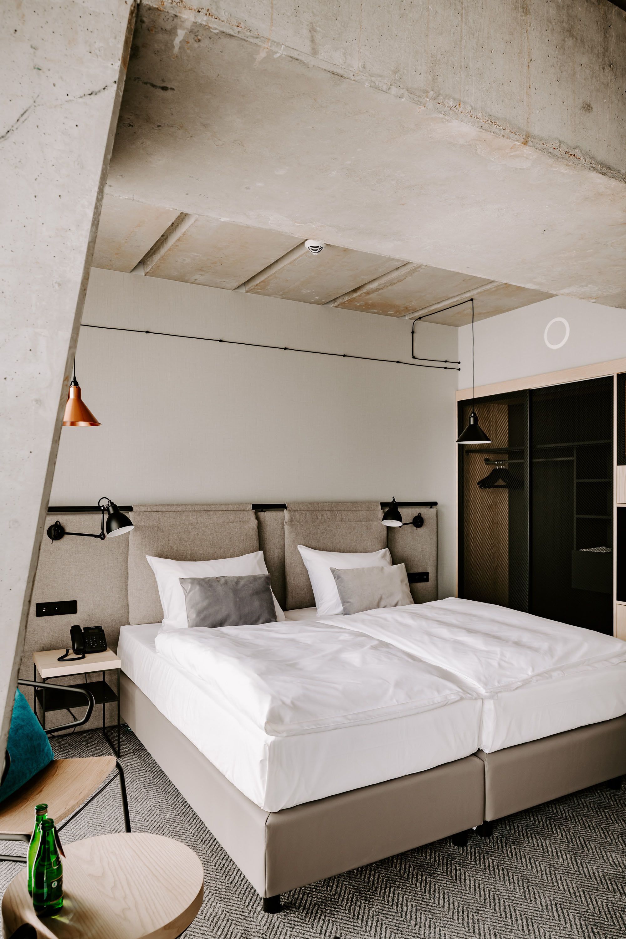 Una habitación de hotel moderna con una cama grande, una mesita de noche, una pequeña mesa redonda con una botella y un armario.