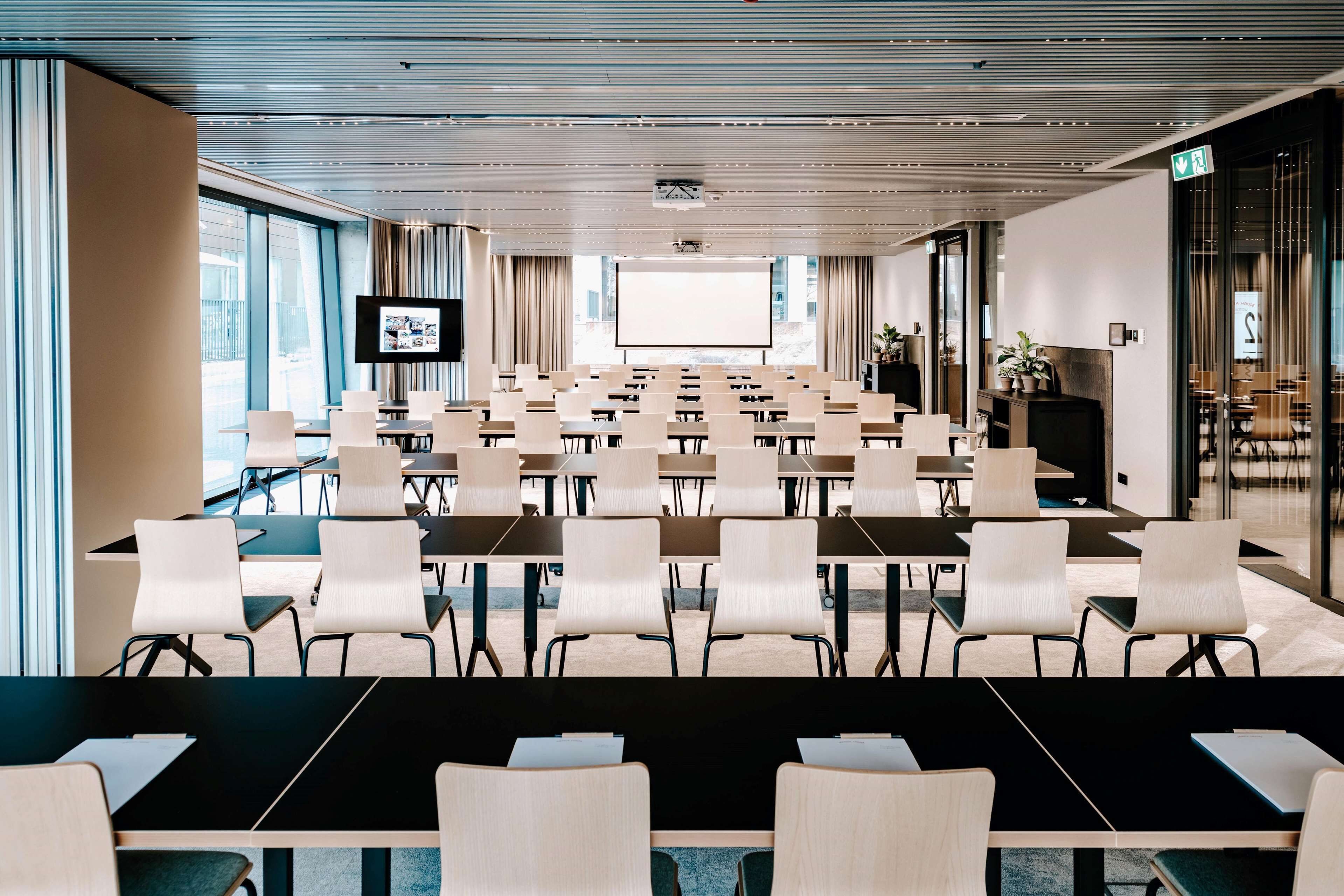 Ein Konferenzraum mit Tischen und Stühlen, einer großen Leinwand an der Wand und großen Fenstern an einer Seite.