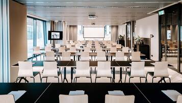 Konferenssitila, jossa on riveissä pöytiä ja tuoleja, suuri näyttö edessä ja suuret ikkunat toisella seinällä.