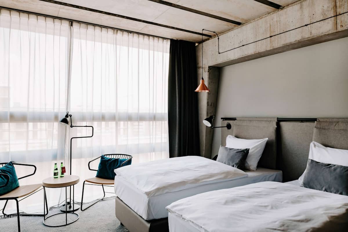 Une chambre d’hôtel avec deux lits, un bureau avec des chaises, une lampe et une vue sur la ville grâce à de grandes fenêtres.