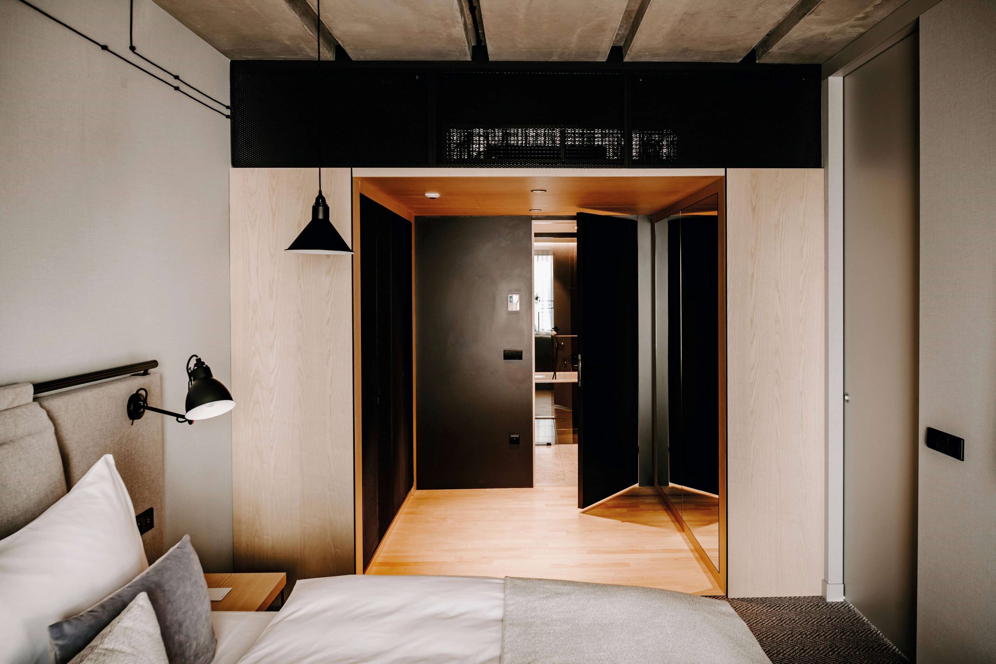 A modern bedroom with a wooden floor, a bed with white bedding, a bedside lamp, and a dark-colored headboard.