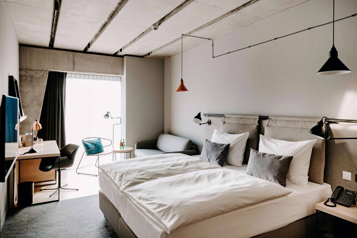 Une chambre d’hôtel moderne avec un grand lit, un bureau et une chaise. On y trouve une fenêtre avec des rideaux, une lampe suspendue et un téléphone sur la table de chevet.