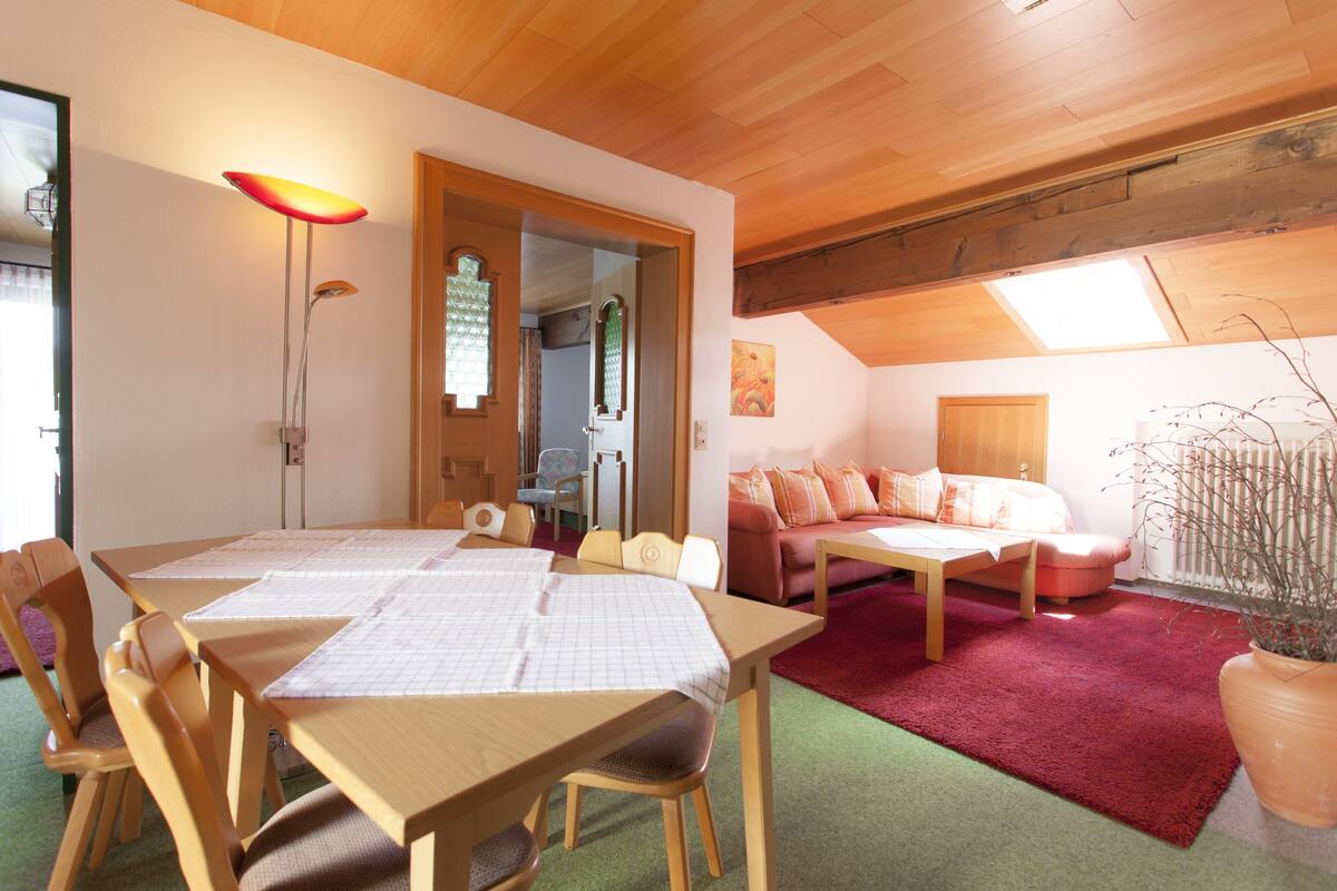 A room with a wooden dining table, chairs, a sofa, and a red carpet.