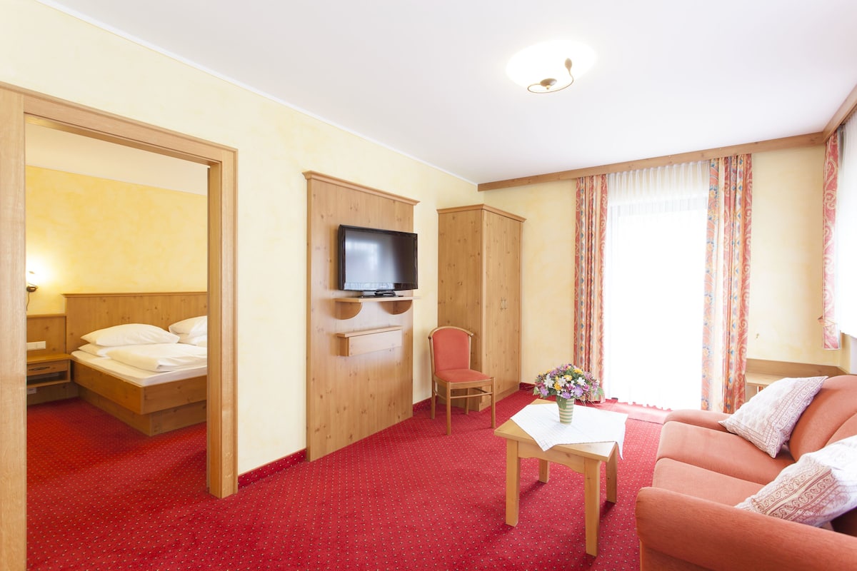 A hotel room with a bed, a TV, a wooden wardrobe, a red carpet, a sofa, and a table with a vase of flowers.