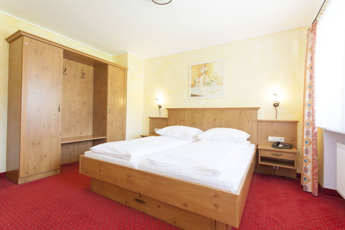 A wooden bed with white linens, a wooden wardrobe, a nightstand with a lamp, and a painting on the wall.