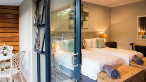 Quarto de hotel com porta de vidro que dá para uma área externa, cama com duas almofadas, mesa de cabeceira e cómoda de madeira.