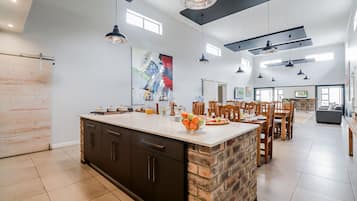Una cocina amplia con una isla grande, armarios oscuros y una pared decorada con ladrillos. Hay varias lámparas colgantes y un cuadro abstracto de gran tamaño en la pared.