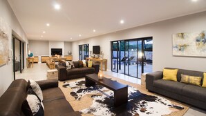 Ampla sala de estar com um sofá grande, uma mesa de centro e uma TV. Há uma área de jantar com uma mesa e cadeiras, e uma varanda com mobiliário para área externa.
